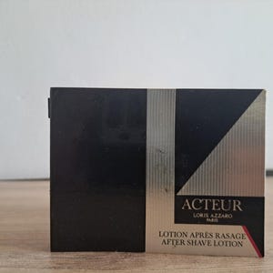 Puede incluir: Caja negra y plateada de loción para después del afeitado ACTEUR Loris Azzaro. La caja tiene un fondo negro con una franja vertical plateada y un diseño diagonal plateado y negro. El texto "ACTEUR" y "AFTER SHAVE LOTION" son visibles.