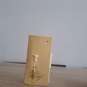 Peut inclure: Un petit flacon de parfum en verre avec un bouchon beige et un liquide clair. Il est placé à côté d'une carte beige avec l'inscription dorée "Davidoff". Le fond est un mur gris clair.