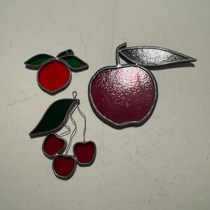 Peut inclure: Trois décorations de fruits en vitrail : une pomme rouge avec une tige et une feuille argentées, une grappe de cerises rouges avec des feuilles vertes et une pêche rouge avec des feuilles vertes. Chaque pièce a une bordure argentée.