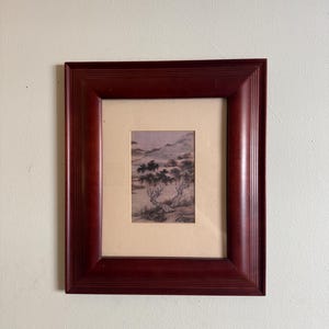 May include: A framed landscape art print depicting trees and mountains in a muted colour palette. The artwork is set within a dark red wooden frame with a layered design. The frame is a rich, dark red.