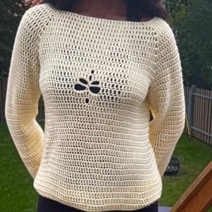 May include: A cream-colored long-sleeve crochet sweater featuring a boat neckline and a central openwork pattern on the chest. The garment is constructed with a consistent horizontal stitch pattern throughout the sleeves and body.
