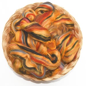 May include: A round woven basket filled with colorful fiber. The fiber is a mix of orange, yellow, red, black, and gray. The basket is made of light brown woven material. The fiber appears soft and fluffy.