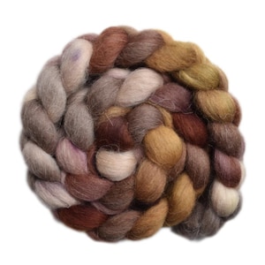 May include: A thick, braided bundle of unspun wool roving featuring a variegated color palette of earthy tones including shades of brown, tan, cream, muted gold, and soft mauve. The fiber has a fuzzy, soft texture throughout the entire coil.