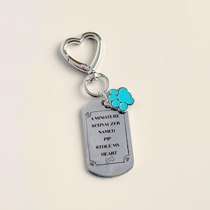 May include: A silver-colored keychain with a heart-shaped clasp and a dog tag. The tag reads "A Miniature Schnauzer Named Pip Stole My Heart." A blue paw print charm hangs from the tag.