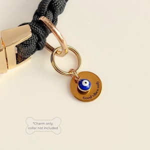 May include: A gold-colored charm with a blue and white evil eye design, attached to a gold ring. The charm is round and has the words "Good vibes only" engraved on it. The charm is attached to a black braided collar with a gold clasp.