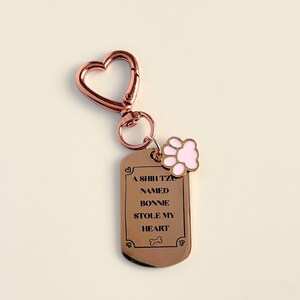 May include: A rose gold keychain with a heart-shaped clasp and a dog tag pendant. The tag reads "A Shih Tzu named Bonnie stole my heart" with a pink paw print charm. A cute accessory for dog lovers.