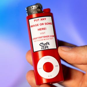 May include: A red cylindrical lighter featuring a white rectangular label with the text PUT ANY IMAGE OR SONG HERE! VISIT CRAFTRIFTSHOP.COM INSTAGRAM @CRAFTRIFT and a white circular control interface with MENU, play, pause, and skip icons.