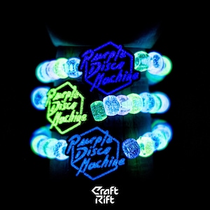 May include: Three glowing bracelets with the text "Purple Disco Machine" in blue and green. The bracelets are made of small, round, glowing beads. The background is black.
