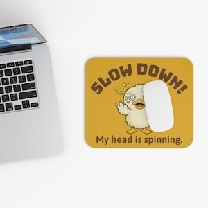 May include: Mustard yellow mousepad with the words "SLOW DOWN!" and a cartoon duck with a dizzy expression. The text "My head is spinning." is below the duck. A white computer mouse rests on the mousepad.