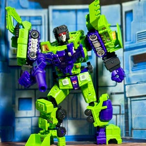 Puede incluir: Una figura de acción verde, morada y negra de un robot con diseño de vehículo de construcción. La figura tiene una cabeza negra con ojos rojos y está posada dinámicamente. Cuenta con una pala y otras piezas de vehículo de construcción.