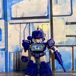 May include: A small blue and grey robot keychain featuring a cassette player chest design with a purple Decepticon logo. The figure has red eyes, a white face mask, and a blue arm-mounted weapon. A metal keyring is attached to the top of the head.
