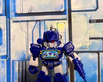 May include: A small blue and grey robot keychain featuring a cassette player chest design with a purple Decepticon logo. The figure has red eyes, a white face mask, and a blue arm-mounted weapon. A metal keyring is attached to the top of the head.