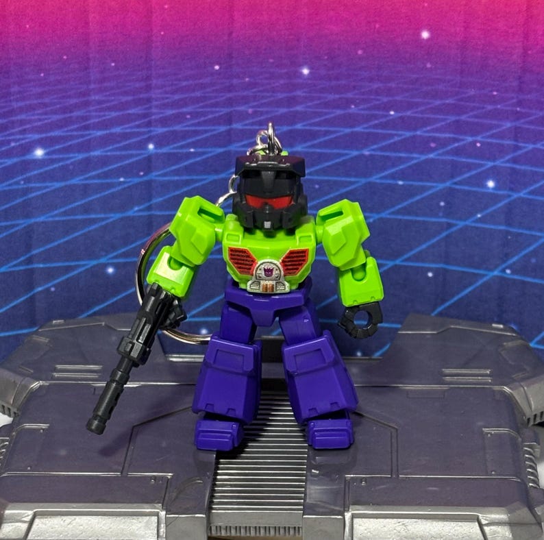 Op de afbeelding: Een kleine robotsleutelhanger met een zwarte helm met rode ogen, een fel limoengroene romp met rode borstopeningen en paarse benen. De robot houdt een zwarte blaster in zijn rechterhand en heeft een klein paars Decepticon-logo op de borst.