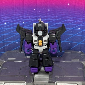 Op de afbeelding: Een kleine, gestileerde robot-actiefiguur sleutelhanger met een zwart, wit en paars kleurenschema. Het figuur heeft een wit gezicht, een centraal borstpaneel met een oranje vlam-achtige afbeelding en beweegbare vleugels aan de schouders.