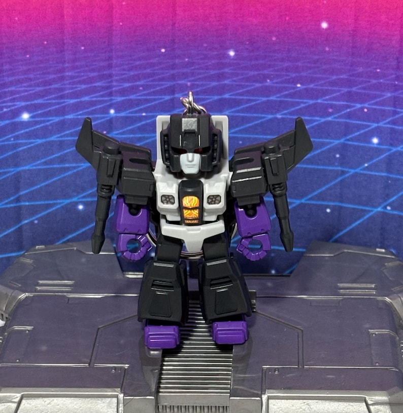 Op de afbeelding: Een kleine, gestileerde robot-actiefiguur sleutelhanger met een zwart, wit en paars kleurenschema. Het figuur heeft een wit gezicht, een centraal borstpaneel met een oranje vlam-achtige afbeelding en beweegbare vleugels aan de schouders.