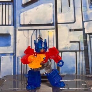 Op de afbeelding: Een doorschijnende Optimus Prime actiefiguur sleutelhanger met een rode romp, blauw hoofd en benen, en een feloranje bijlaccessoire in de rechterhand. Het figuur heeft een zilveren Autobot-insigne op de linkerschouder en een metalen ring.