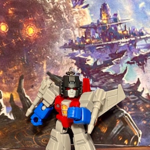 Op de afbeelding: Een kleine, gestileerde robot-actiefiguur sleutelhanger in een grijs, rood en blauw kleurenschema. Het figuur heeft een zwarte helm, rode ogen en een wit gezicht, met beweegbare ledematen en vleugels aan de achterkant, voorzien van een metalen ring.