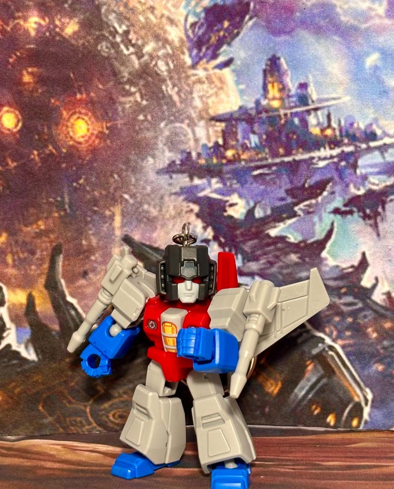 Op de afbeelding: Een kleine, gestileerde robot-actiefiguur sleutelhanger in een grijs, rood en blauw kleurenschema. Het figuur heeft een zwarte helm, rode ogen en een wit gezicht, met beweegbare ledematen en vleugels aan de achterkant, voorzien van een metalen ring.