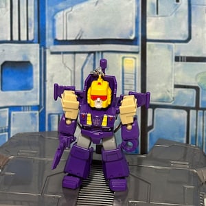 Op de afbeelding: Een kleine paars-gele robot actiefiguur sleutelhanger met een gele kop, rood vizier en wit gezicht. De robot heeft beige schoudervullingen, een paarse romp met gele accenten en een paars Decepticon-logo op het borstgedeelte.