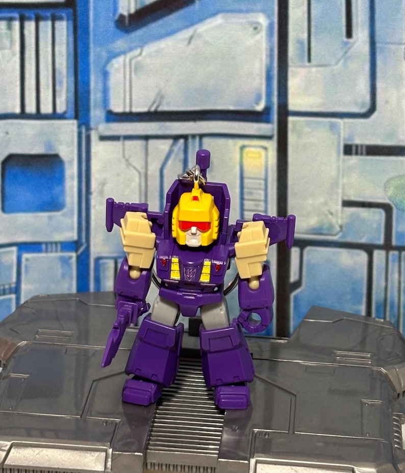 Op de afbeelding: Een kleine paars-gele robot actiefiguur sleutelhanger met een gele kop, rood vizier en wit gezicht. De robot heeft beige schoudervullingen, een paarse romp met gele accenten en een paars Decepticon-logo op het borstgedeelte.