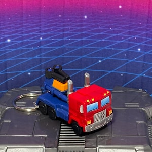 Peut inclure: Un jouet de camion rouge, bleu et jaune avec un porte-clés. La cabine est rouge avec des cheminées argentées et des fenêtres bleues. La remorque est bleue avec une section jaune et des canons noirs. Le jouet est posé sur une surface grise.