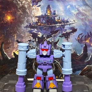 Op de afbeelding: Een paarse en lichtblauwe robot actiefiguur sleutelhanger met een rood vizier, wit gezicht en twee grote cilindrische armbevestigingen. De robot heeft gele driehoekige details op de onderbenen en staat op een metaalgrijs oppervlak.