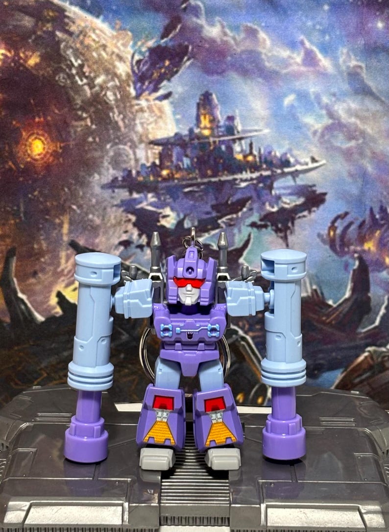 Op de afbeelding: Een paarse en lichtblauwe robot actiefiguur sleutelhanger met een rood vizier, wit gezicht en twee grote cilindrische armbevestigingen. De robot heeft gele driehoekige details op de onderbenen en staat op een metaalgrijs oppervlak.