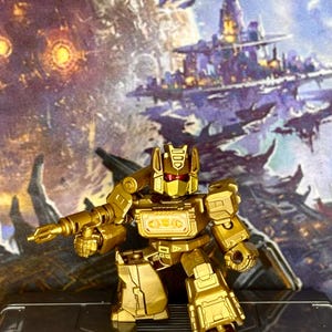 Op de afbeelding: Een metallic gouden robotactiefiguur met rode vizierogen en een borstcompartiment met een Decepticon-embleem. Het figuur houdt een bijpassende gouden blaster in de rechterhand en staat in een stijve, rechtopstaande houding met beweegbare ledematen.