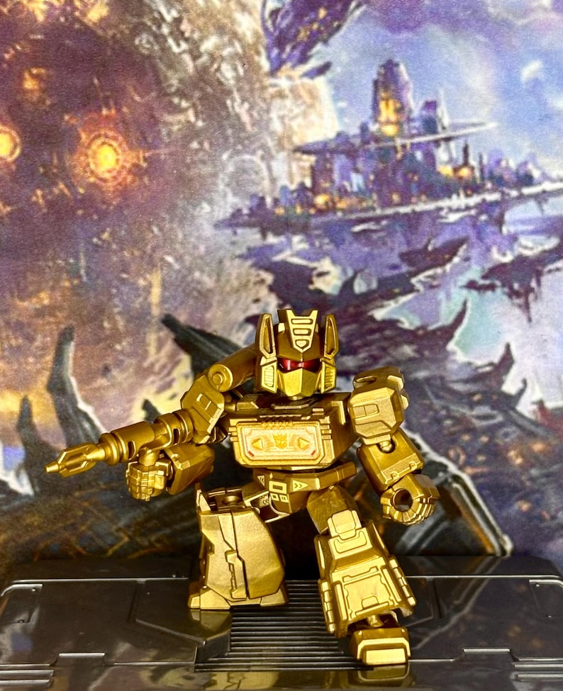 Op de afbeelding: Een metallic gouden robotactiefiguur met rode vizierogen en een borstcompartiment met een Decepticon-embleem. Het figuur houdt een bijpassende gouden blaster in de rechterhand en staat in een stijve, rechtopstaande houding met beweegbare ledematen.