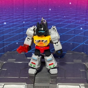 Op de afbeelding: Een kleine robot actiefiguur sleutelhanger met een zwarte kop en blauw vizier, een gele borstplaat met oranje en zwarte details, een rode taille en grijze ledematen. Het figuur heeft een doorschijnend rood wapen aan de rechterarm.
