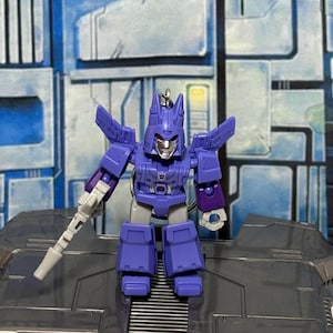 Op de afbeelding: Een paarse en grijze robot actiefiguur sleutelhanger met een puntige helm, een zilverkleurig gezicht en een klein Decepticon-embleem op de borst. Het figuur heeft beweegbare armen, een wit handwapen en een metalen ring aan de bovenkant.