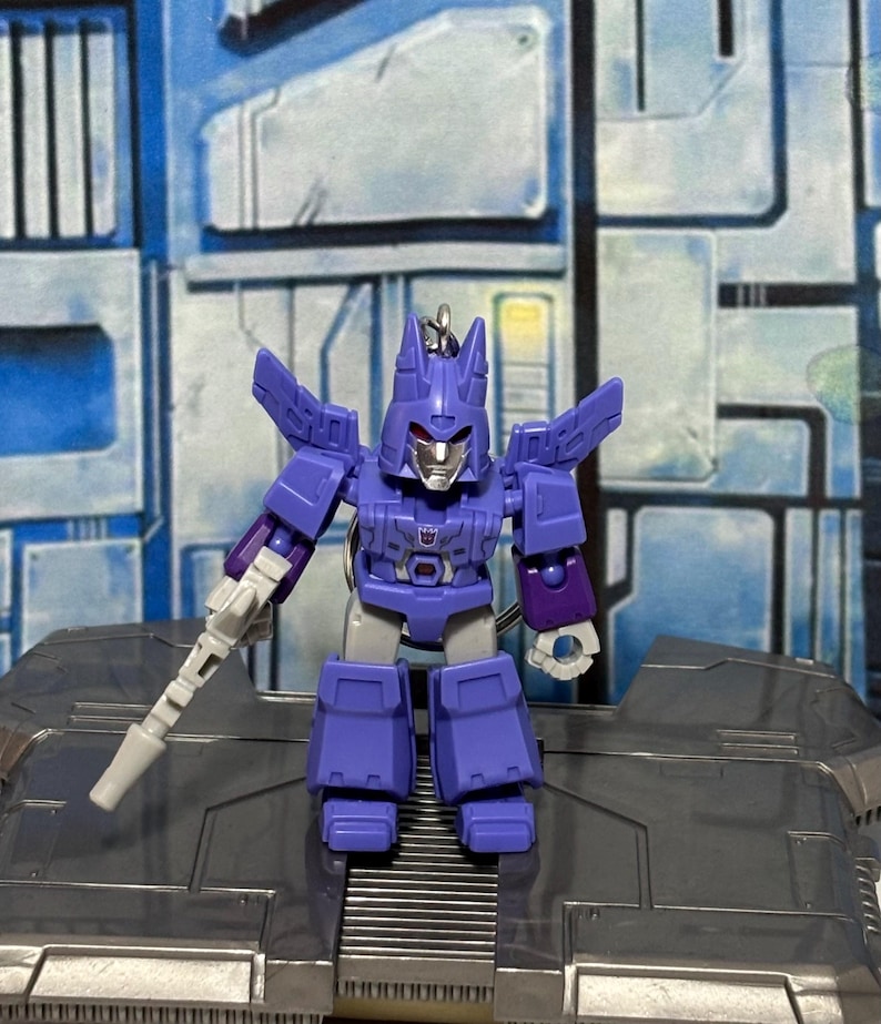 Op de afbeelding: Een paarse en grijze robot actiefiguur sleutelhanger met een puntige helm, een zilverkleurig gezicht en een klein Decepticon-embleem op de borst. Het figuur heeft beweegbare armen, een wit handwapen en een metalen ring aan de bovenkant.