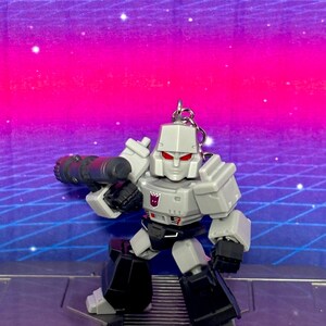 Op de afbeelding: Een grijze en zwarte Megatron actiefiguur sleutelhanger met een Decepticon-embleem op de borst, rode ogen en een groot kanon op de arm. Het personage staat in een rechtopstaande houding met hoekige, robotachtige pantserdetails en een metalen ring.