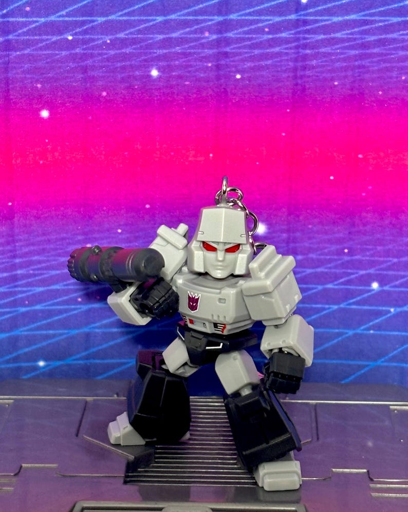 Op de afbeelding: Een grijze en zwarte Megatron actiefiguur sleutelhanger met een Decepticon-embleem op de borst, rode ogen en een groot kanon op de arm. Het personage staat in een rechtopstaande houding met hoekige, robotachtige pantserdetails en een metalen ring.