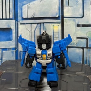 Op de afbeelding: Een kleine, beweegbare robot-actiefiguur sleutelhanger met een blauw, zwart en lichtgrijs kleurenschema. Het figuur heeft een hoekig hoofd, vleugels aan de schouders en een centraal borststuk met een geel accent. Een metalen ring is aan de bovenkant van het hoofd bevestigd.