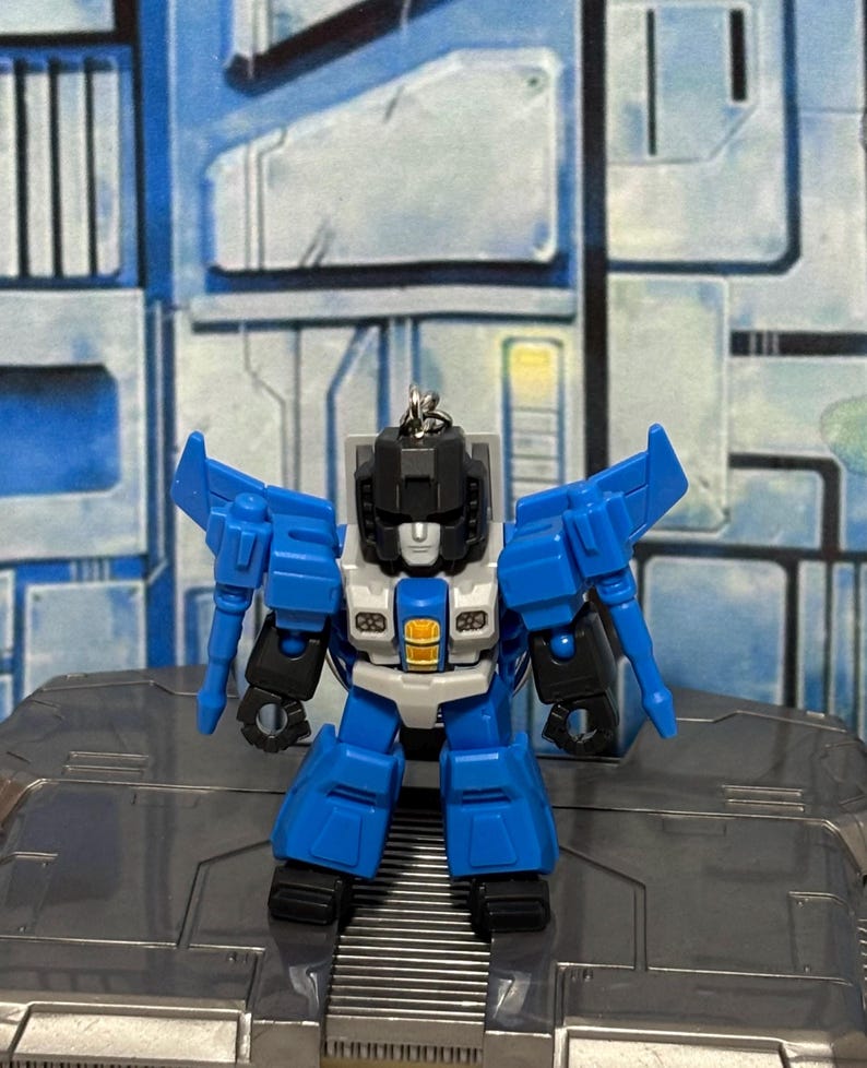 Op de afbeelding: Een kleine, beweegbare robot-actiefiguur sleutelhanger met een blauw, zwart en lichtgrijs kleurenschema. Het figuur heeft een hoekig hoofd, vleugels aan de schouders en een centraal borststuk met een geel accent. Een metalen ring is aan de bovenkant van het hoofd bevestigd.