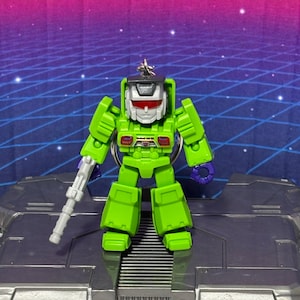Op de afbeelding: Een fel limoengroene robotsleutelhanger met een zilveren helm met rood vizier, paarse handen en een grijs blaster-accessoire in de rechterhand. Het figuurtje heeft een kleine metalen ring aan de bovenkant van het hoofd om op te hangen.