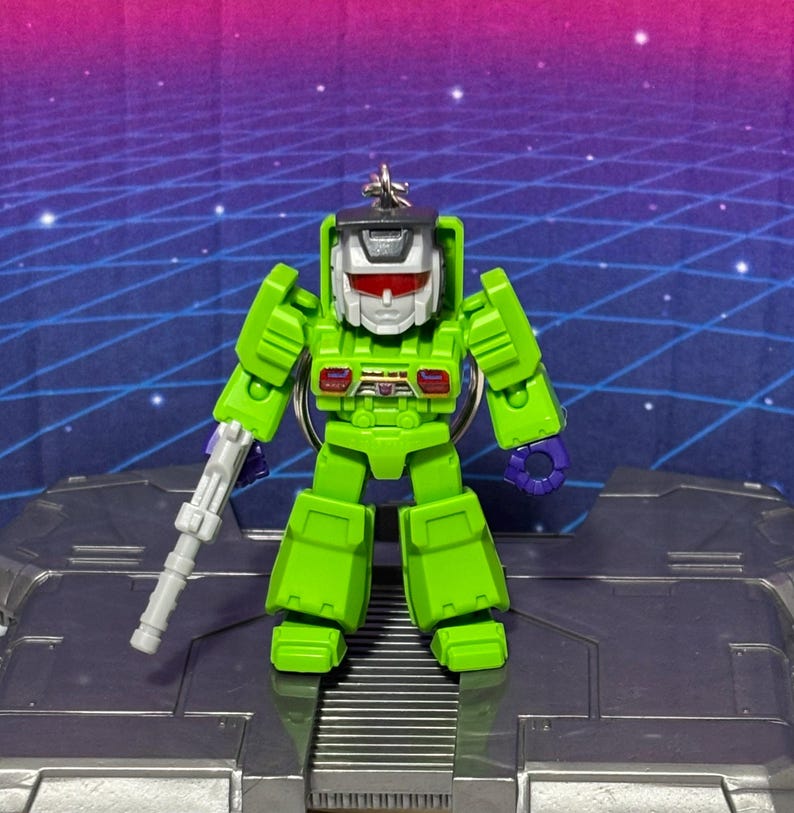 Op de afbeelding: Een fel limoengroene robotsleutelhanger met een zilveren helm met rood vizier, paarse handen en een grijs blaster-accessoire in de rechterhand. Het figuurtje heeft een kleine metalen ring aan de bovenkant van het hoofd om op te hangen.