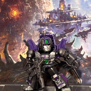 Op de afbeelding: Een kleine, beweegbare robotactiefiguur sleutelhanger met een paars en zwart kleurenschema en zilveren accenten. Het figuur heeft een metallic zilveren gezicht, een paarse helm met een groen detail op het voorhoofd en houdt een donkergrijs geweer vast.