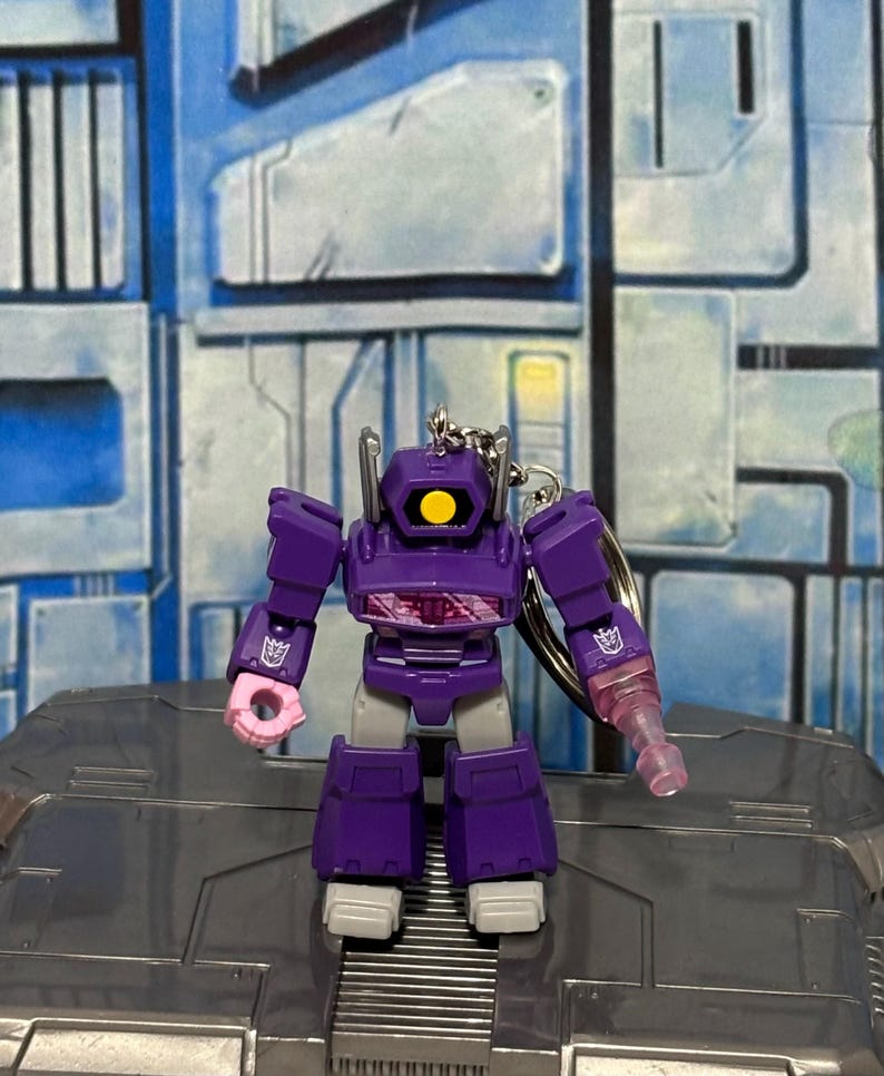 Op de afbeelding: Een paarse en grijze robot-sleutelhanger met &eacute;&eacute;n geel oog, een doorschijnend roze armopzetstuk en een Decepticon-logo op elke schouder. De robot heeft een hoekig, mechanisch ontwerp met een zilverkleurige metalen ring aan de bovenkant.