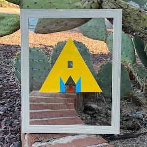 May include: A framed artwork featuring a yellow triangle with a square cutout and blue accents. The artwork is set against a backdrop of a brick wall and a cactus, with a light-colored wooden frame.
