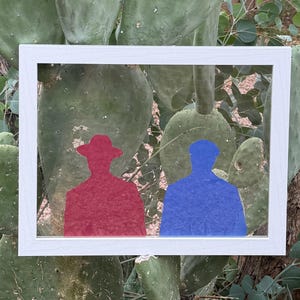 May include: A framed artwork featuring two silhouetted figures against a backdrop of cacti. One figure is red and wearing a hat, the other is blue. The frame is white.