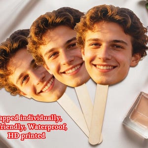 May include: Three face cutouts on wooden sticks, each depicting a smiling person with light brown hair. The cutouts are arranged in a fan shape. The text "Wrapped individually, Eco-friendly, Waterproof, HD printed" is visible.