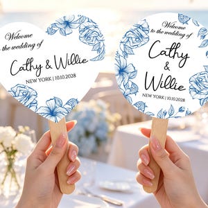 Puede incluir: Dos abanicos de boda con mangos de madera, uno en forma de corazón y otro circular. Ambos tienen fondos blancos, dibujos florales en azul y el texto negro Welcome to the wedding of Cathy & Willie, NEW YORK | 10.10.2028.