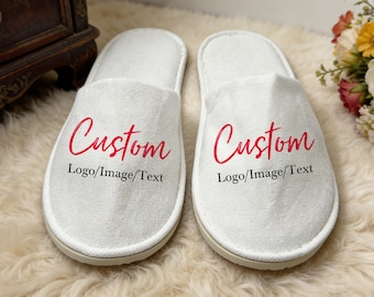 May include: A pair of white open-toe slippers featuring the word Custom in a red script font, with the text Logo/Image/Text printed in black underneath on each slipper. The slippers have a textured fabric surface and a white trim.