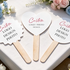 Op de afbeelding: Drie witte handwaaiers met houten handvatten, met een geschulpte rand, een hartvorm en een cirkelvorm. Elke waaier toont de rode tekst Custom en zwarte tekst met LOGO DESIGN PHOTO of LOGO / PHOTO DESIGN.