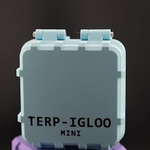 Puede incluir: Una caja TERP-IGLOO MINI azul claro con tapa con bisagras, colocada sobre una caja morada. El texto "TERP-IGLOO MINI" está impreso en negro en la parte delantera de la caja azul claro. Las cajas tienen un diseño cuadrado.