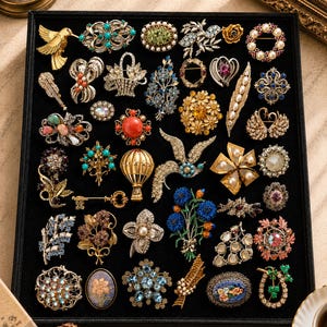 Può includere: Una collezione di spille in metallo assortite disposte su un vassoio espositore nero. Le spille presentano vari disegni tra cui uccelli, fiori, chiavi, mongolfiere e forme geometriche, arricchite da strass colorati, perle e smalto.