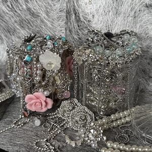 May include: Two ornate, silver-toned containers overflowing with jewelry. The jewelry includes necklaces, bracelets, rings, and brooches, featuring pearls, turquoise, and clear crystals. Decorative pink and white floral accents are also visible.