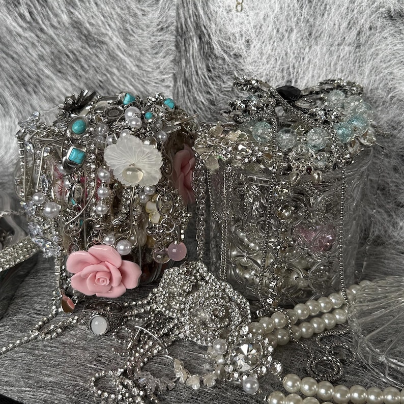 May include: Two ornate, silver-toned containers overflowing with jewelry. The jewelry includes necklaces, bracelets, rings, and brooches, featuring pearls, turquoise, and clear crystals. Decorative pink and white floral accents are also visible.